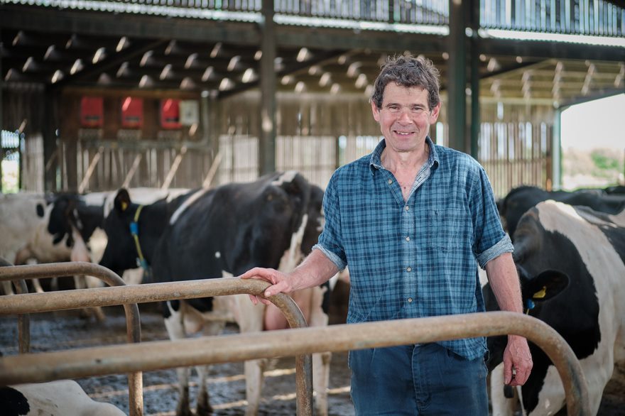 Dairy farmer in check shirt stood holding onto a cubicle with cows in the background